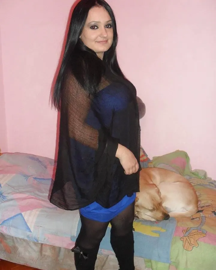 Femme aux cheveux noirs portant une robe bleue sous une écharpe noire transparente, debout près d'un lit rose avec un chien endormi.