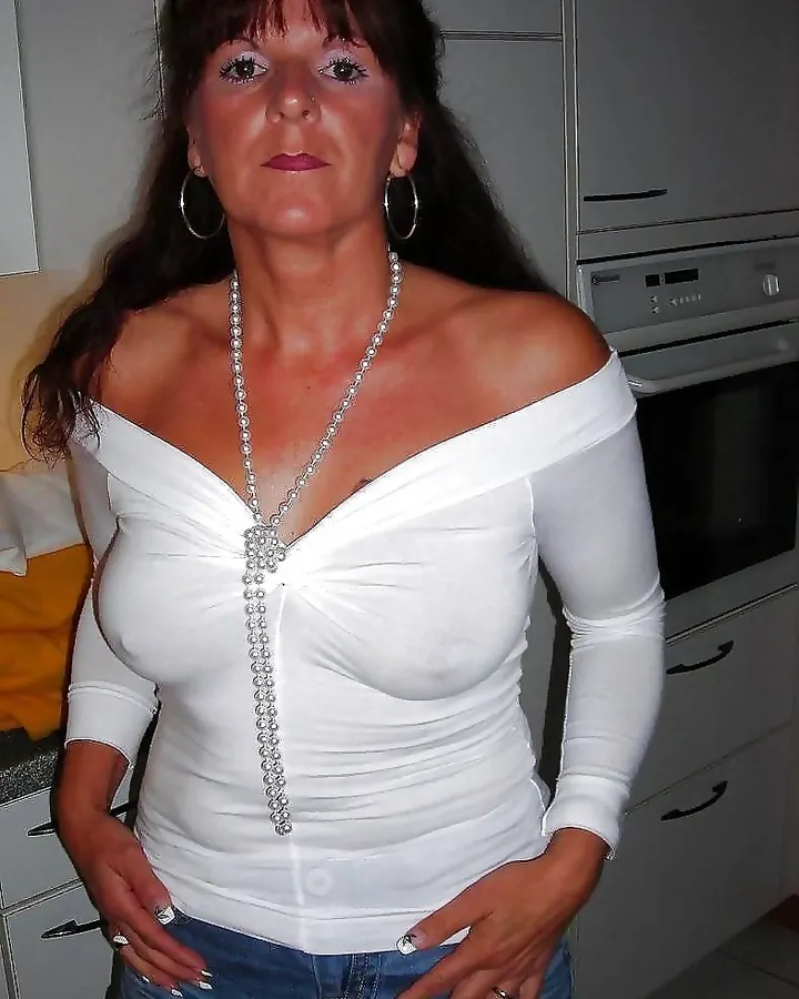 Femme aux cheveux bruns foncés, portant un haut blanc décolleté et un collier de perles, debout dans une cuisine moderne, mains sur les hanches.