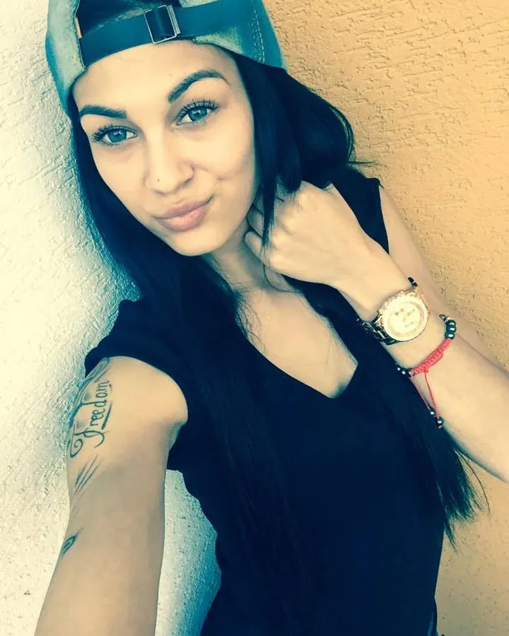 Femme latino aux cheveux bruns-noirs, portant un haut noir et un string noir, assise sur un canapé en cuir, regardant vers l'appareil.