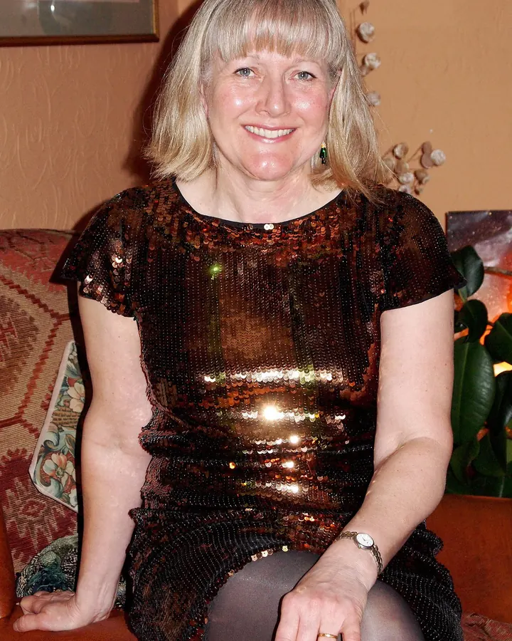Femme aux cheveux blancs courts, portant une robe scintillante marron, assise sur un canapé rouge.