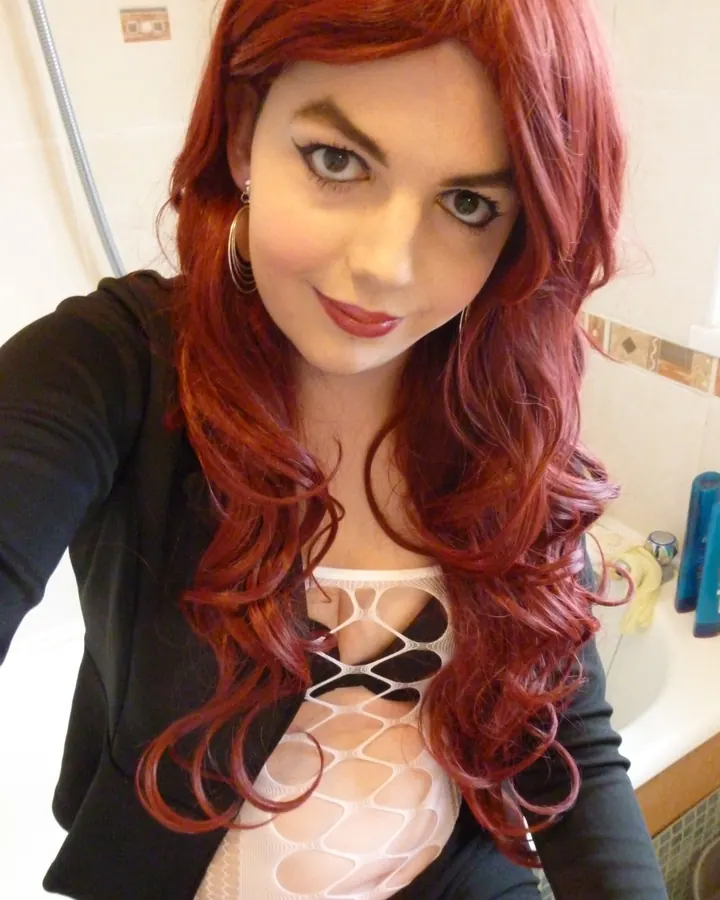 Femme aux cheveux roux mi-longs, portant un haut noir, un pantalon noir, un corset noir avec des brides croisées, debout contre un mur blanc.
