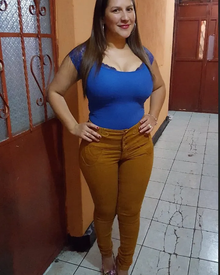 Femme aux cheveux châtains et yeux marrons en haut bleu et pantalon marron, posant dans un couloir