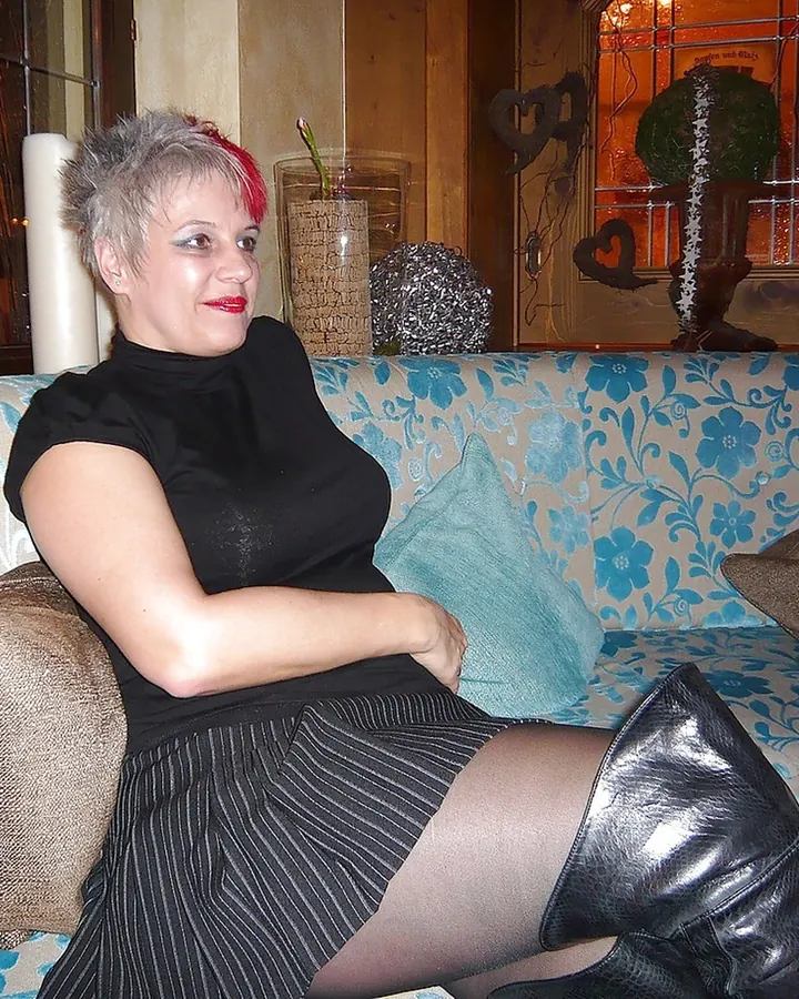 Femme aux cheveux gris portant une robe rouge, debout dans un salon avec un canapé bleu
