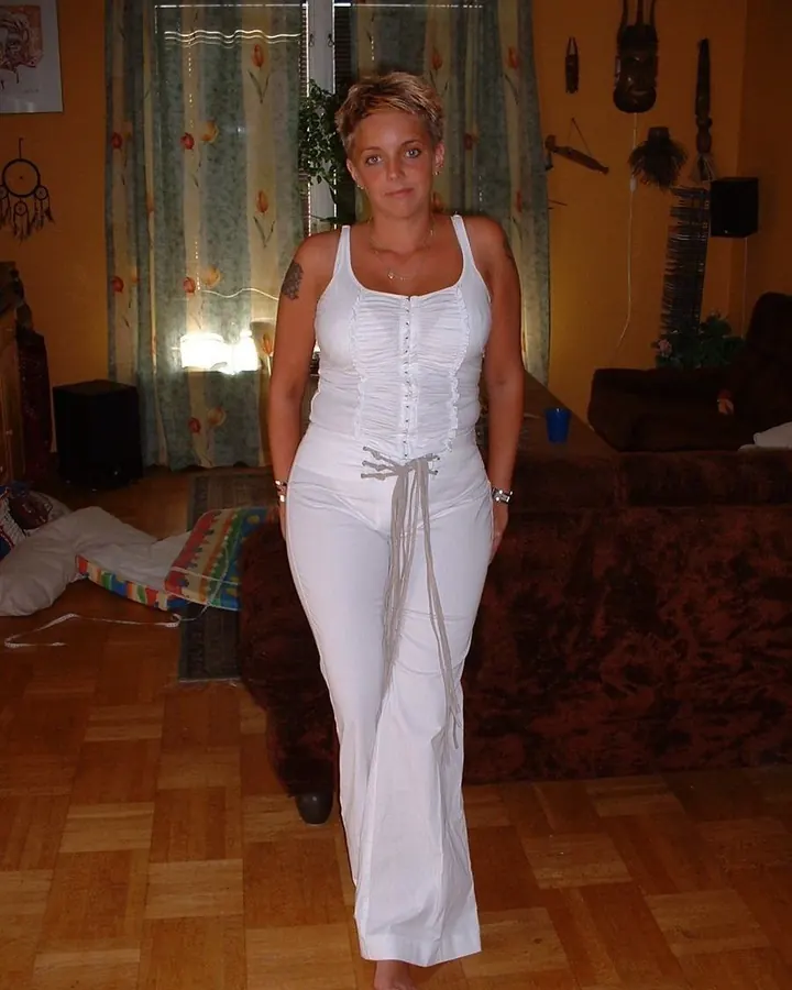 Femme aux cheveux courts blonds portant une tenue blanche, debout dans un salon avec des rideaux colorés