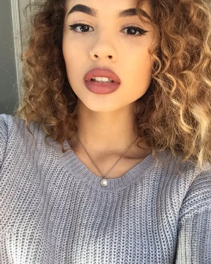 Jeune femme aux cheveux blonds et yeux marrons, prenant un selfie, portant un pull gris et un piercing au nez.