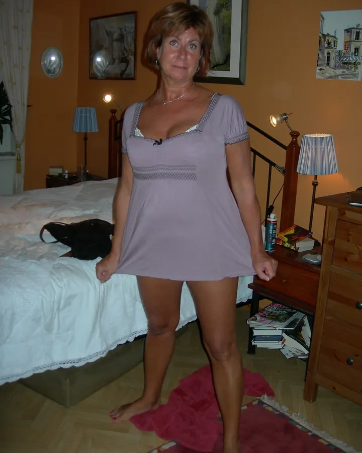 Femme aux cheveux roux et yeux bleus, debout dans une chambre, portant une robe lilas courte.