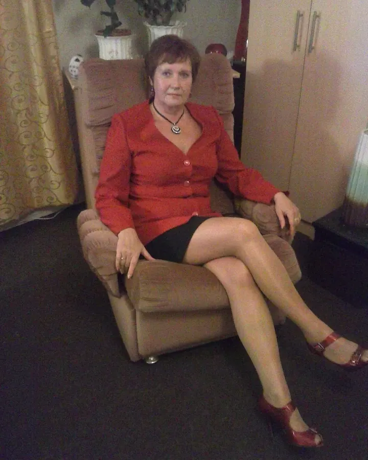 Femme plus âgée aux cheveux châtains assise dans un fauteuil, vêtue d'une chemise rouge et d'une jupe noire.