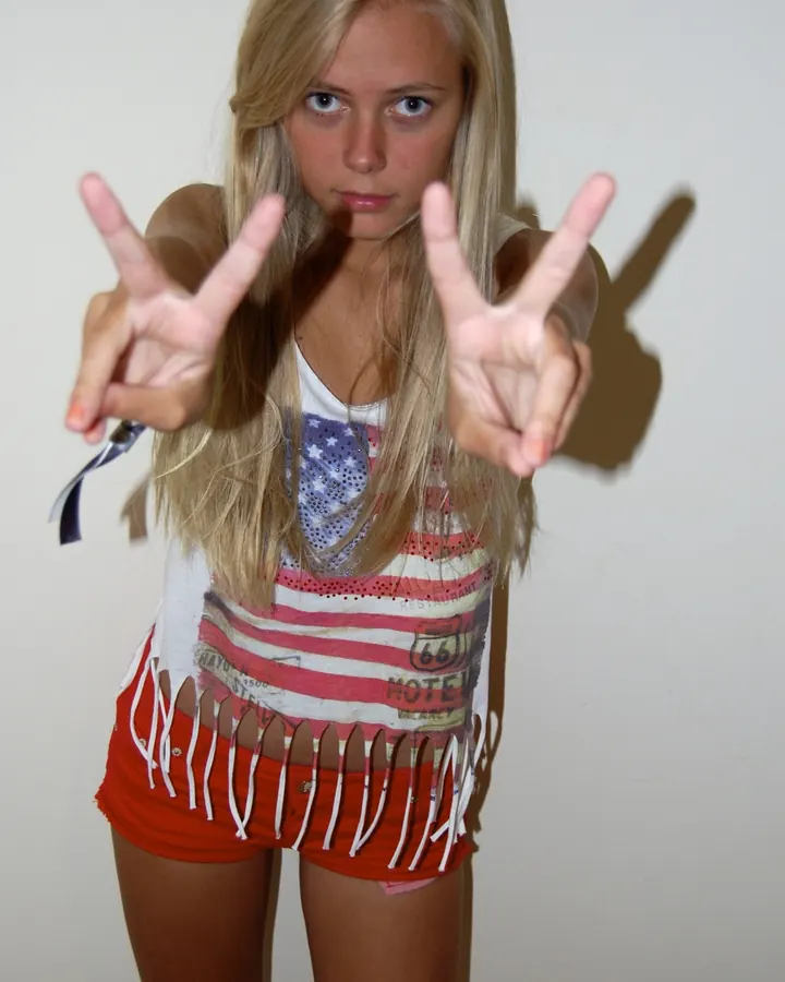 Jeune femme blonde aux yeux bleus faisant un signe de paix avec les deux mains, vêtue d'un haut américain et d'un short rouge.