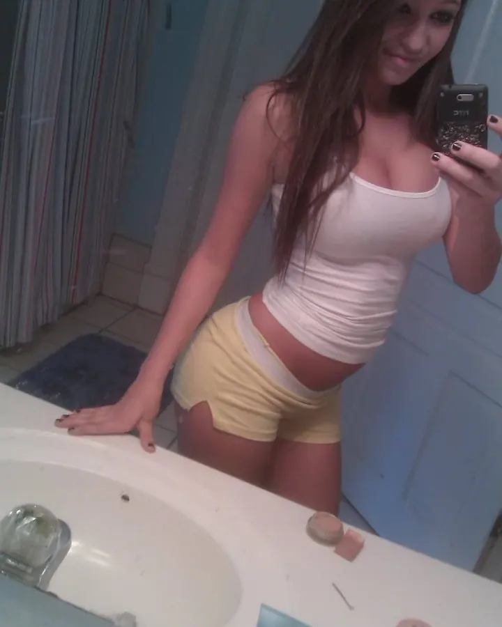 Jeune femme aux cheveux châtains prenant un selfie dans une salle de bain, vêtue d'un haut blanc et d'un short jaune.