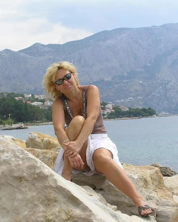 Femme blonde en robe à motifs camouflage, posant près d'un canapé marron, main sur la hanche.