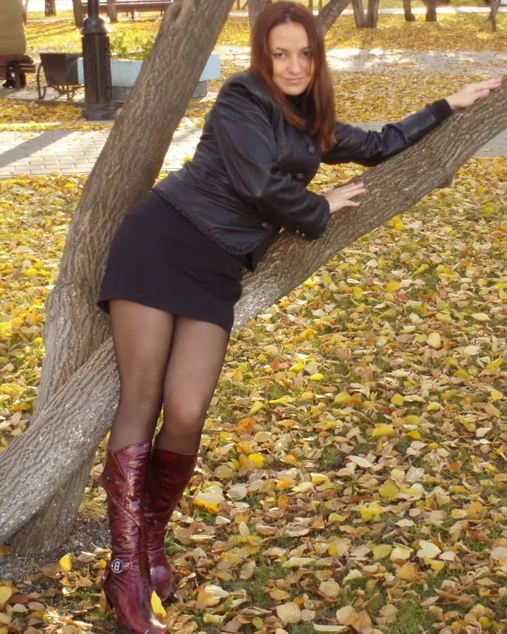 Femme rousse en tenue sexy noire, chaussures à talons, posant près d'un pilier intérieur.