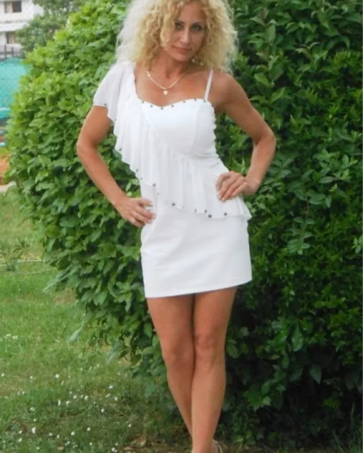 Femme aux cheveux platine en robe blanche, debout devant un buisson vert