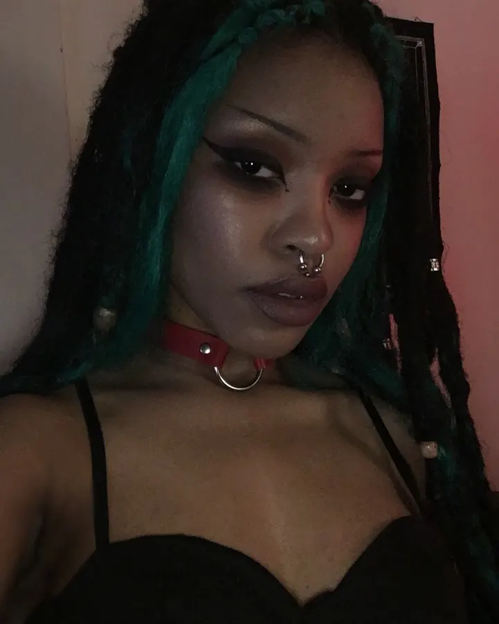 Femme africaine aux cheveux tressés verts, collier rouge, regard intense, vêtue d'une tenue noire dans un cadre sombre.