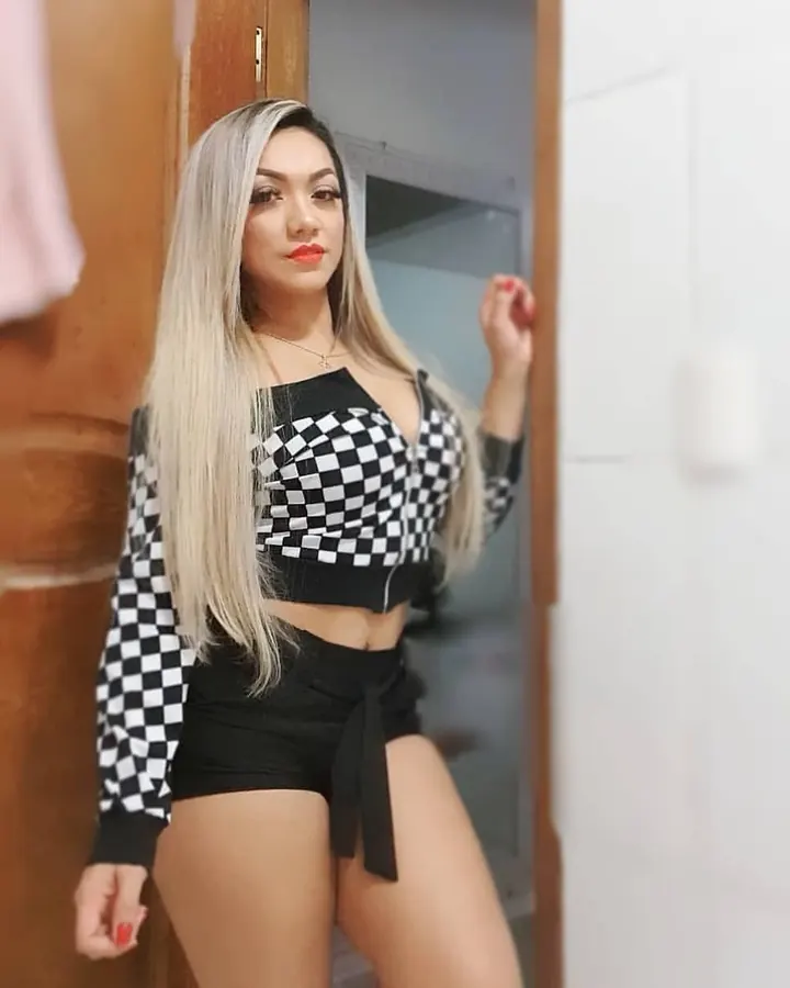 Femme aux cheveux blonds, crop-top à carreaux noir et blanc, shorts noirs, appuyée contre une porte en bois.