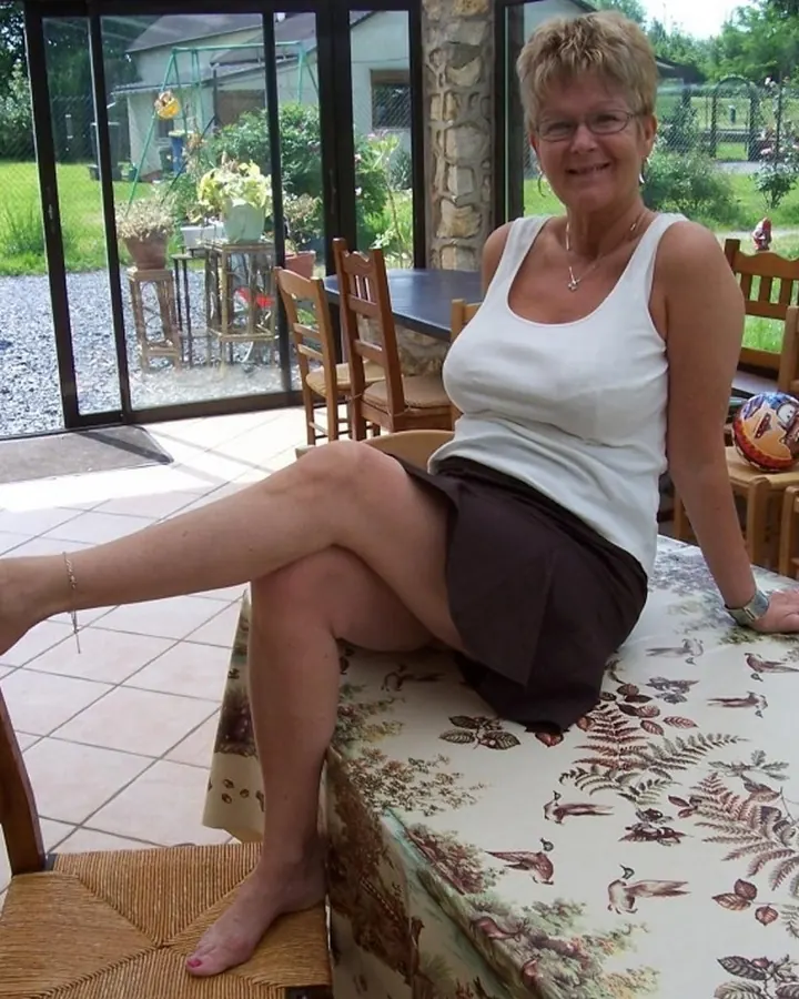 Femme blonde aux cheveux courts, débardeur blanc, jupe courte marron, assise sur une table de jardin, pied posé sur une chaise.