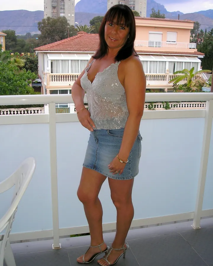 Femme latine aux cheveux bruns, debout sur un balcon, portant un haut gris transparent et une jupe en jean, avec des sandales à talons.