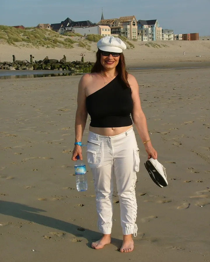 Femme mûre aux cheveux roux, debout sur une plage, portant un haut noir asymétrique, un pantalon blanc, un chapeau blanc et des lunettes de soleil, tenant une bouteille d'eau et une chaussure.