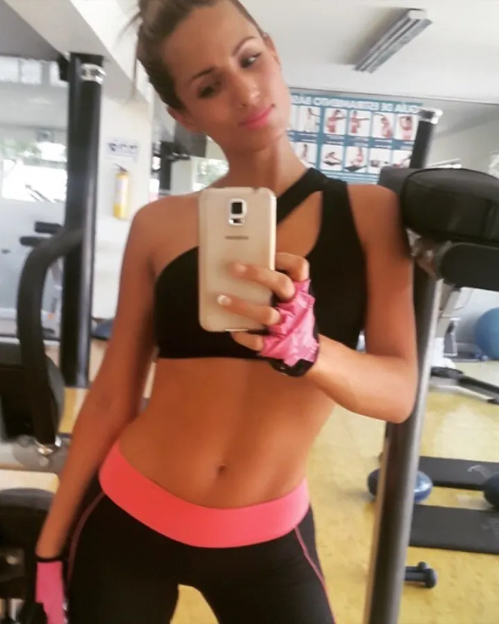 Femme latine aux cheveux bruns, prenant un selfie dans un gymnase, portant un haut de sport noir et un legging noir avec une bande rose.