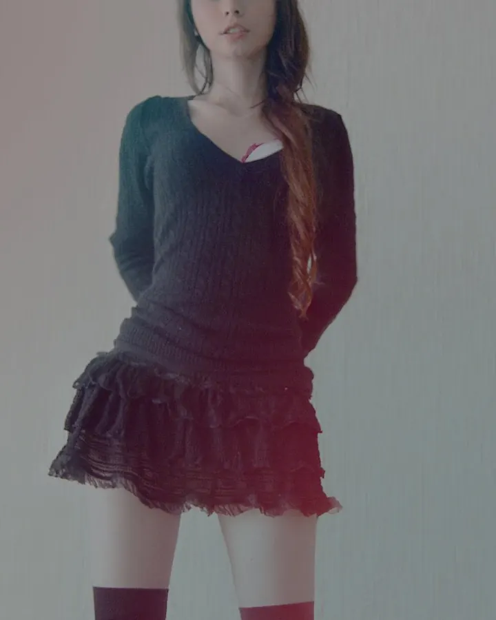 Jeune femme aux cheveux bruns portant un pull noir et une jupe noire à volants, avec des collants foncés et des bottes