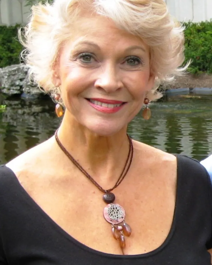 Femme souriante aux cheveux blonds courts, collier en bois, robe noire, devant un étang et un jardin verdoyant