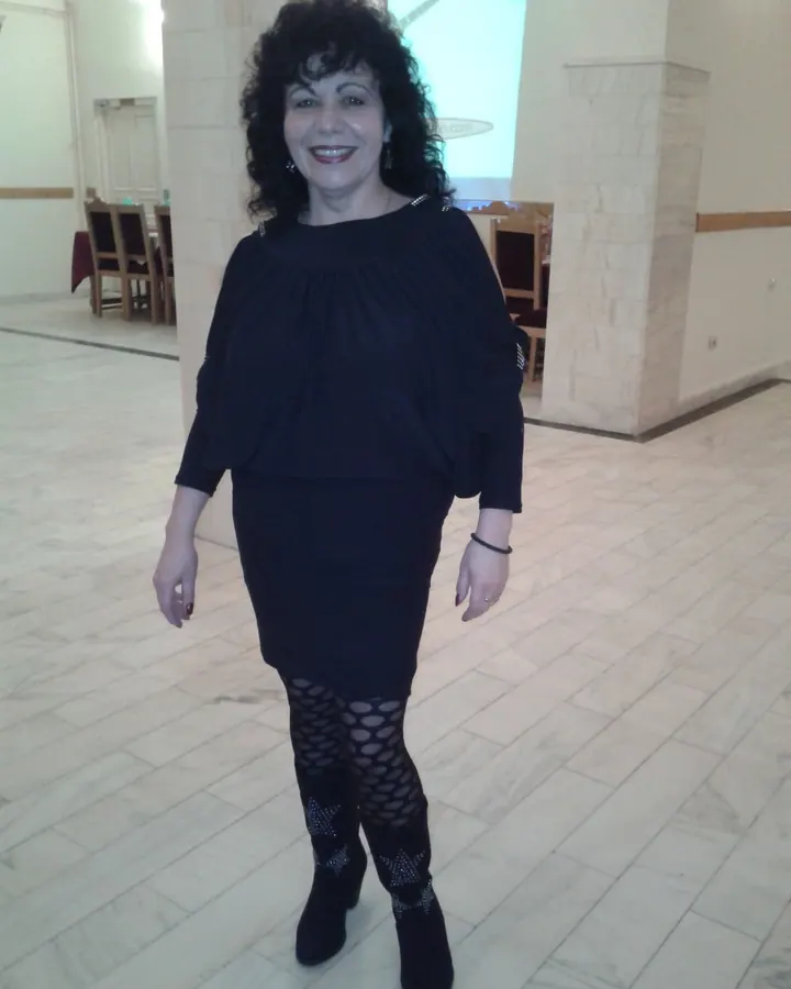 Femme aux cheveux noirs courts, en robe noire, bottes noires, collants à motifs, souriante dans une salle de réception