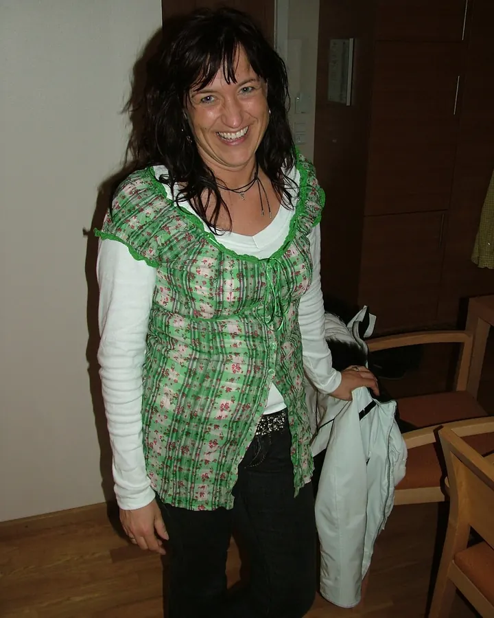 Femme brune souriante, portant un haut vert à motifs, debout près d’un mur en pierre avec un sac blanc à côté.