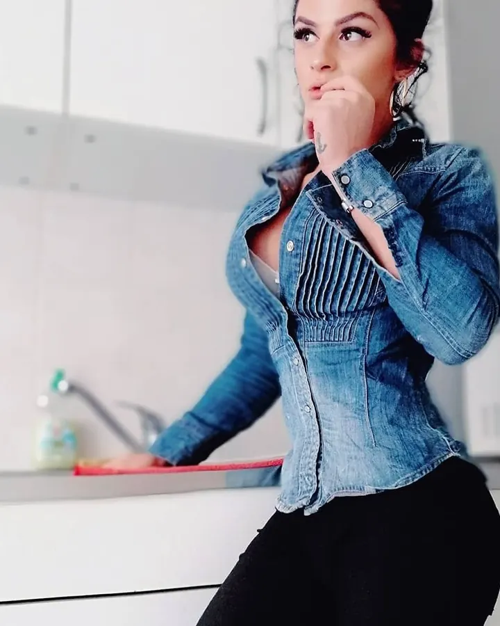 Femme aux cheveux noirs, portant une chemise en jean bleue, posant dans une cuisine blanche avec un bras sur le comptoir.