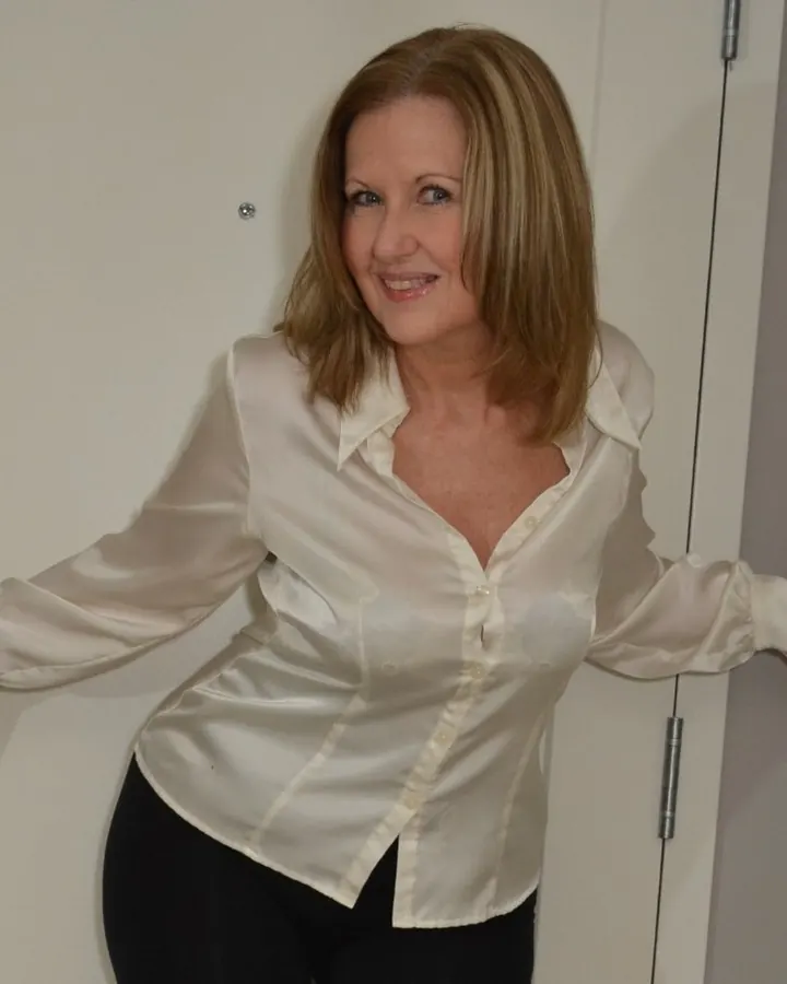 Femme aux cheveux blonds, portant une chemise blanche et une jupe noire, assise sur une chaise en bois avec des bas noirs et des chaussures à talons.