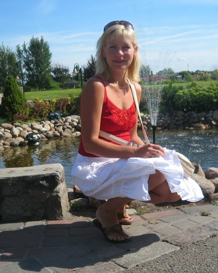 Femme aux cheveux blonds platine, portant une robe rouge et une jupe blanche, assise près d'une fontaine dans un jardin.