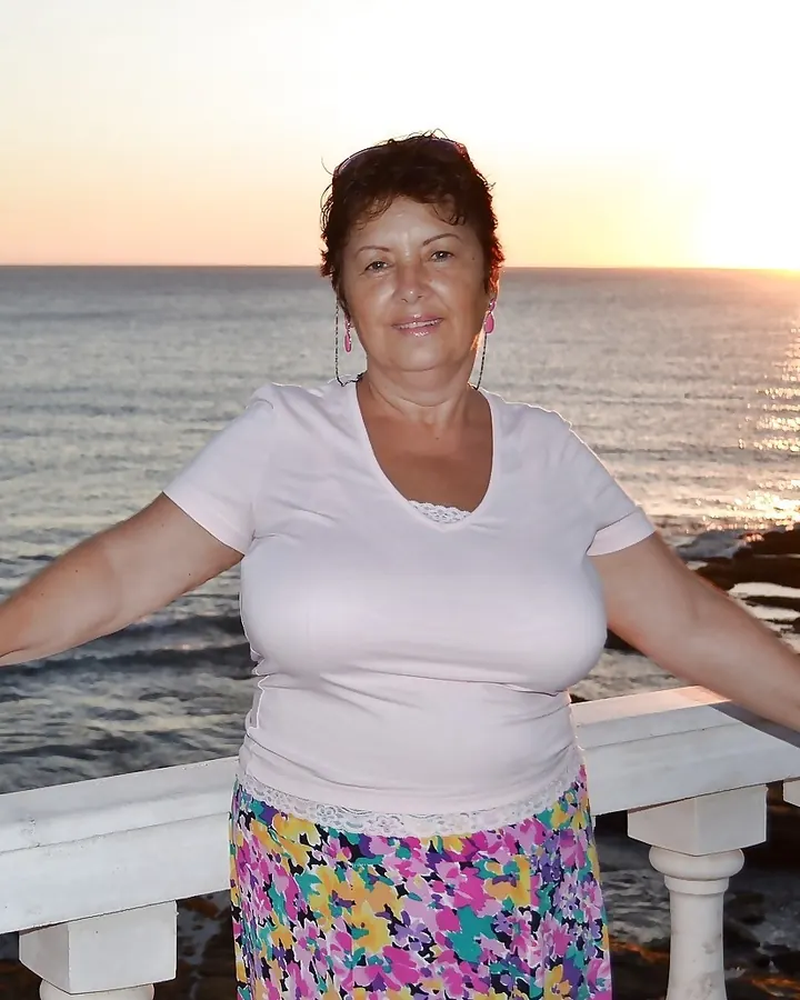 Femme mûre aux cheveux châtains, souriante, en tenue blanche, posant sur une terrasse au coucher du soleil.