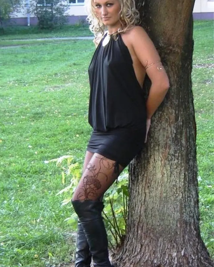 Femme blonde aux cheveux courts, en robe noire, appuyée contre un arbre en extérieur.