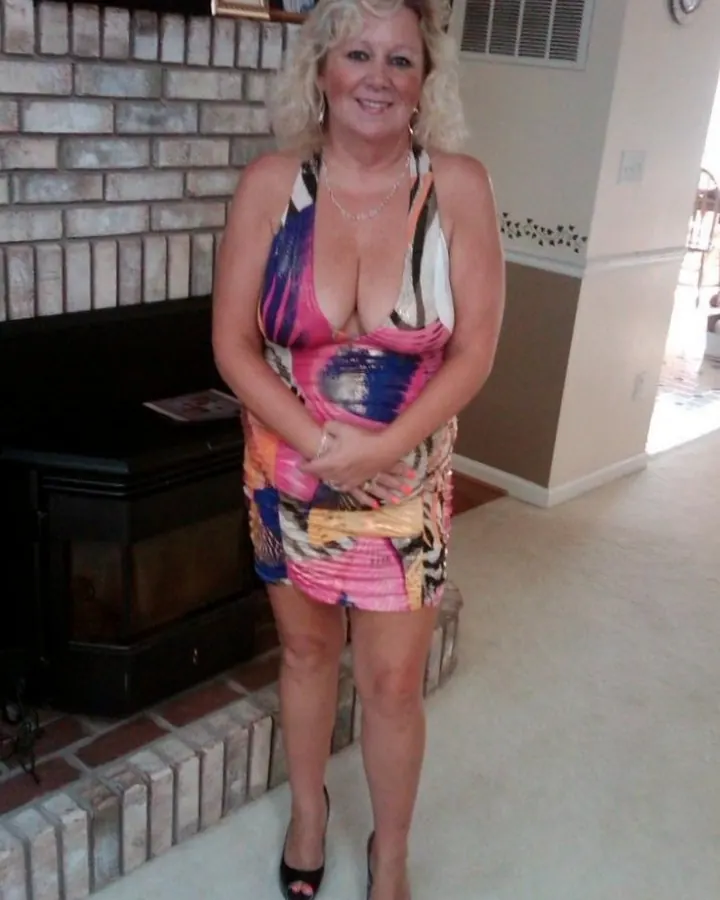 Femme blonde aux cheveux bouclés, souriante, en robe colorée, debout devant une cheminée en briques.