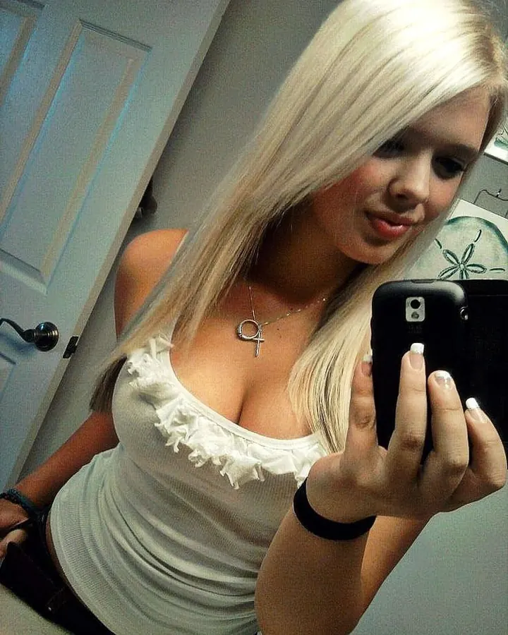 Femme aux cheveux platine en débardeur blanc, prenant un selfie dans un miroir avec un téléphone noir.