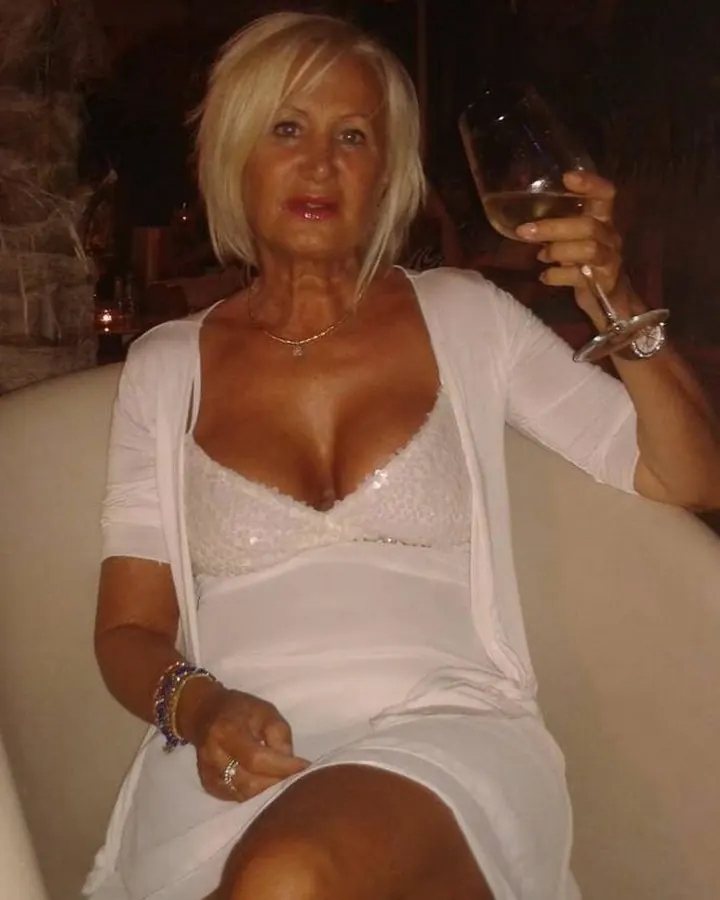 Femme blonde portant une robe blanche et une veste blanche, tenant un verre de vin, assise sur un canapé dans un cadre sombre.