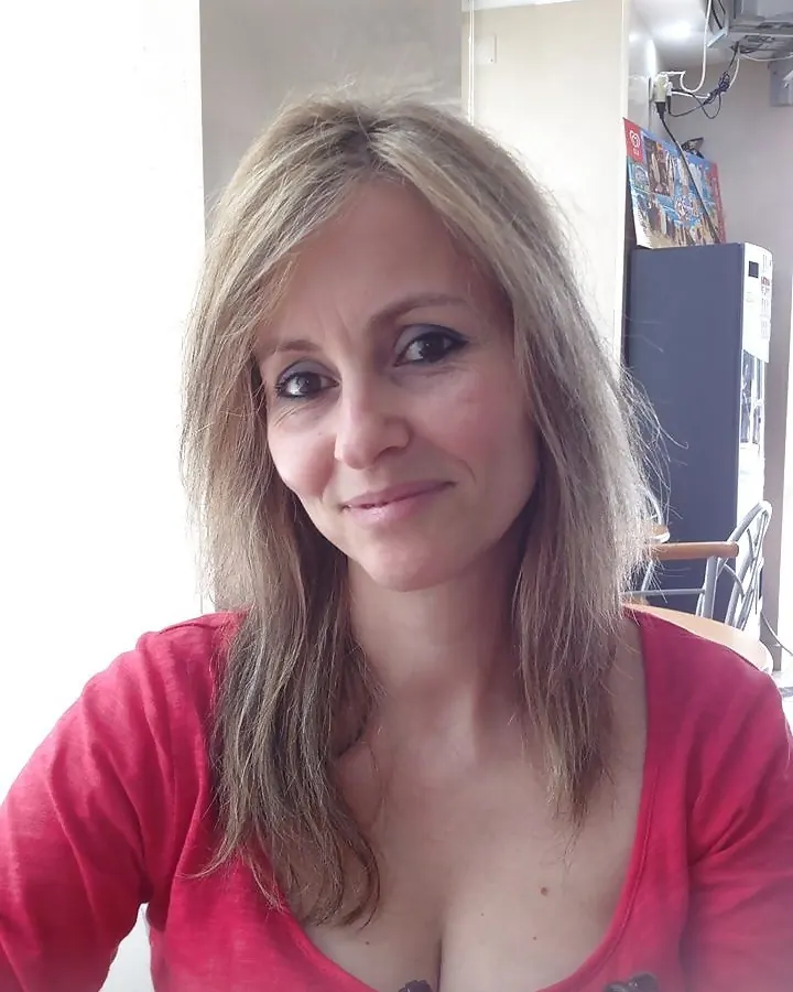 Femme aux cheveux blonds clairs portant un haut rouge décolleté, souriant doucement à la caméra dans un intérieur lumineux.