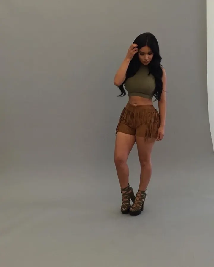 Femme aux cheveux noirs portant un haut vert olive et une jupe courte marron à franges, avec des bottes à talons.