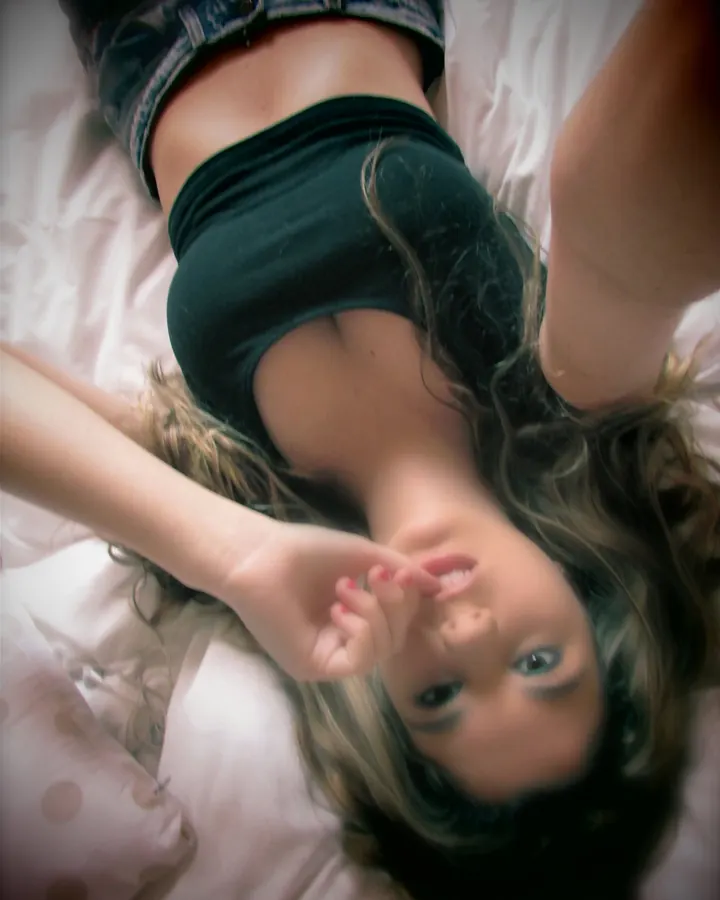Femme aux cheveux noirs portant un haut noir, allongée sur un lit rose, regardant vers le haut.