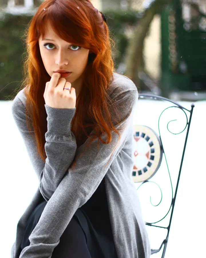 Femme blonde aux yeux bleus, en robe grise, posant sur un lit avec un sourire charmeur.