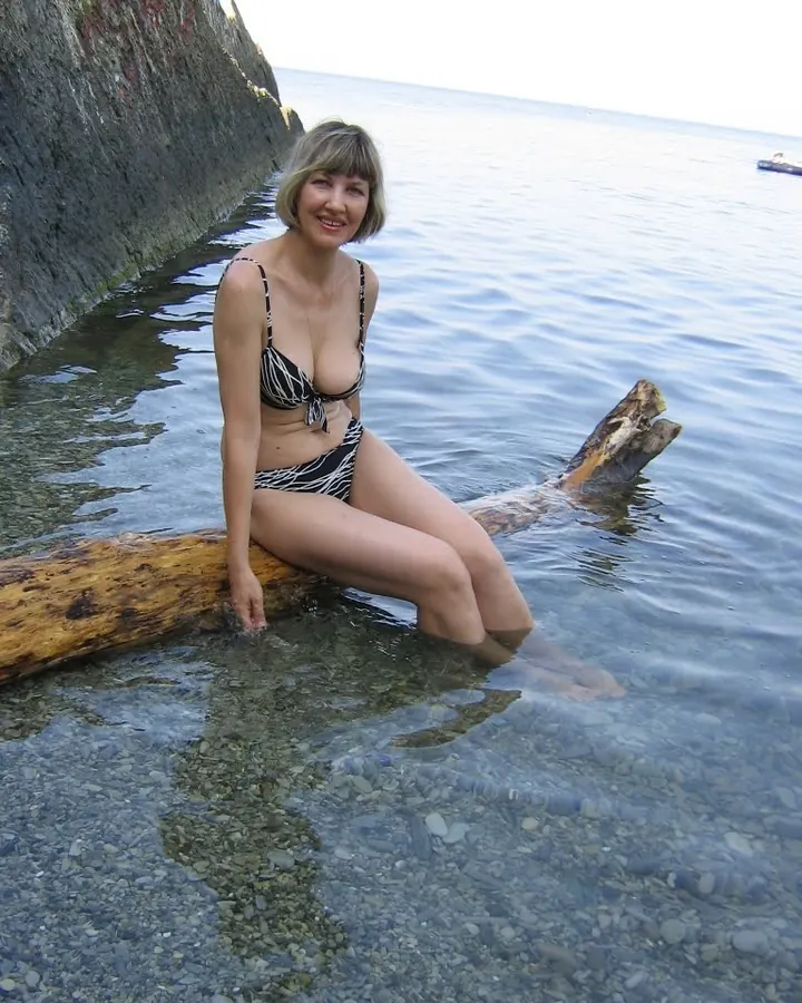 Femme blonde aux yeux marrons, en bikini noir et blanc, assise sur un tronc dans l'eau claire.