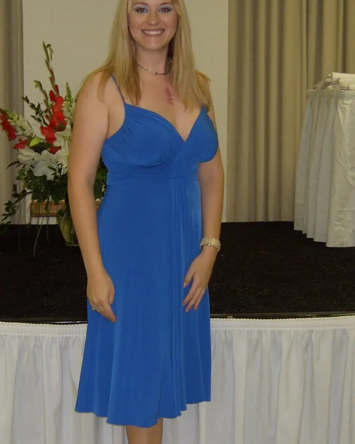 Femme blonde aux yeux bleus, en robe grise, posant debout devant un canapé en cuir avec une main sur la hanche.