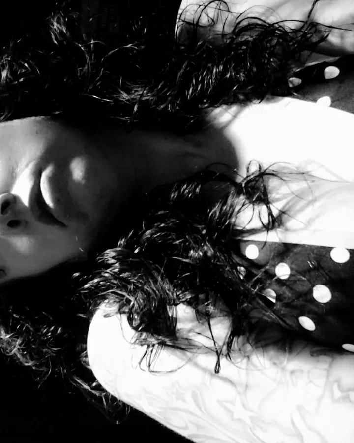 Femme aux cheveux noirs en noir et blanc, allongée, regardant vers le haut, vêtue d’un haut à pois, tatouage visible.
