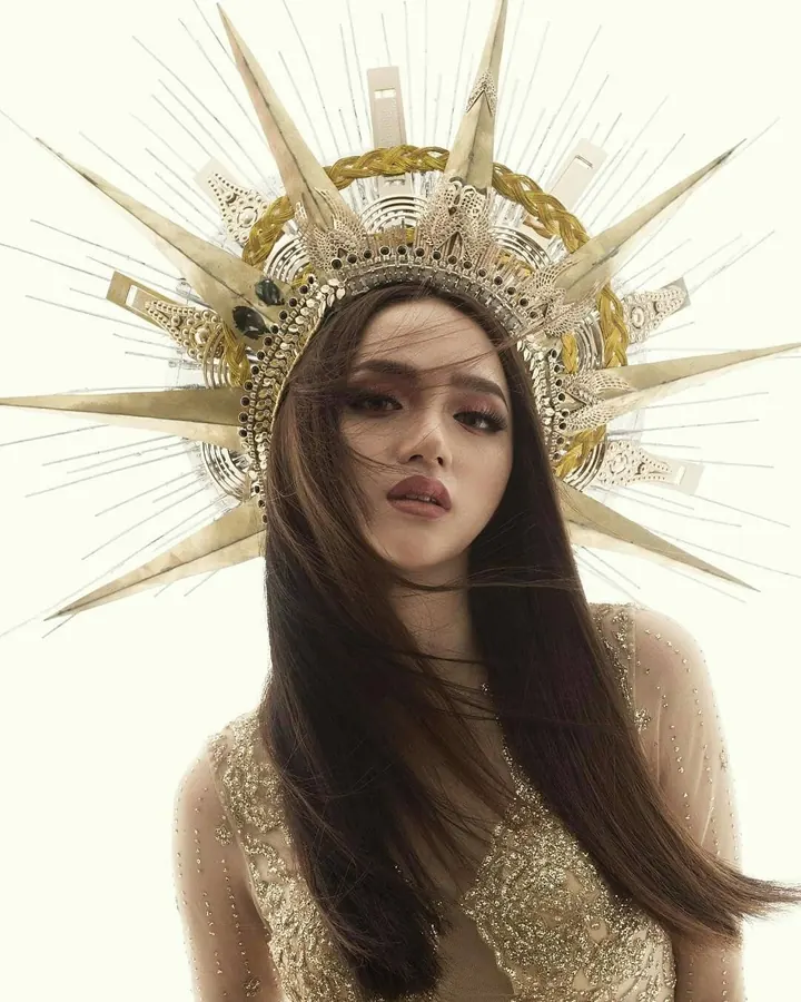 Femme aux cheveux bruns portant une robe dorée et une couronne dorée en forme de soleil, regardant vers l'avant.