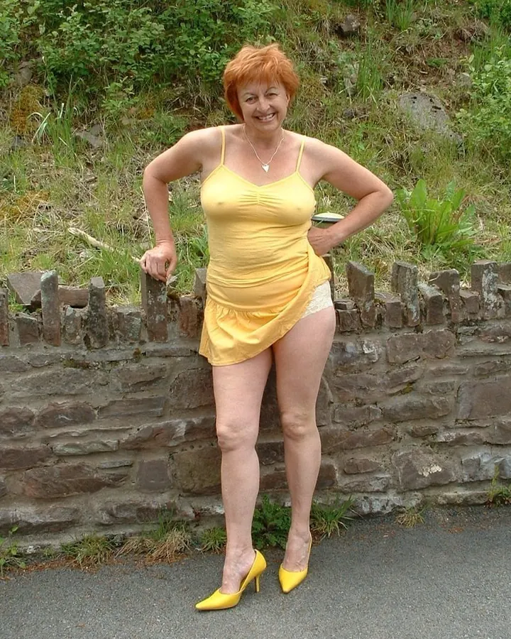 Femme aux cheveux roux, en robe jaune courte et chaussures jaunes, pose contre un mur de pierre en extérieur.