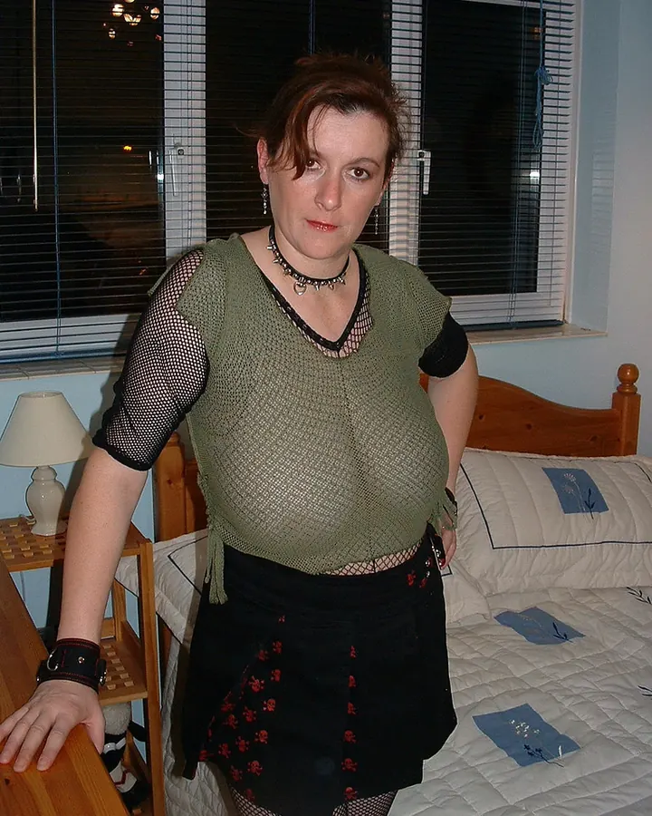 Femme européenne en haut vert olive à maille et jupe noire, pose dans une chambre près d'un lit.