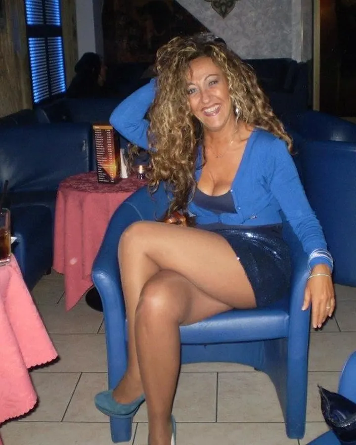Femme aux cheveux blonds bouclés, souriante, portant un haut bleu et une jupe courte bleue pailletée, assise sur un fauteuil bleu dans un bar.