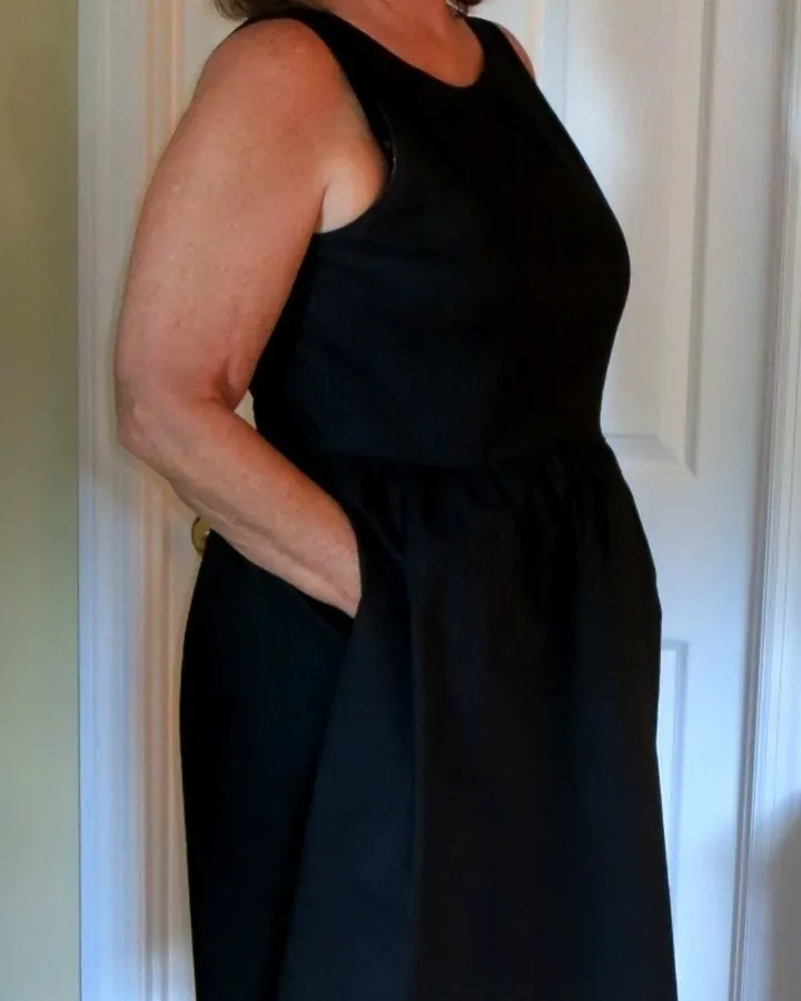 Femme blonde aux cheveux courts, souriante, portant une robe noire en satin avec des rubans rouges, assise sur un canapé marron.