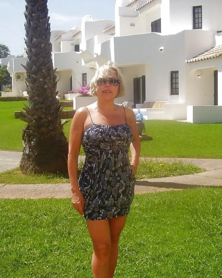 Femme blonde aux cheveux courts, portant des lunettes de soleil, debout sur une pelouse verte devant des bâtiments blancs.