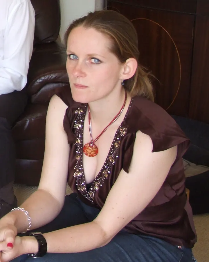 Femme aux cheveux blonds, portant un haut marron orné de strass, assise sur un canapé en cuir marron.