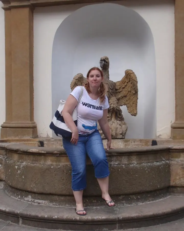 Femme assise sur un banc de pierre, devant une fontaine, en t-shirt blanc et jeans.