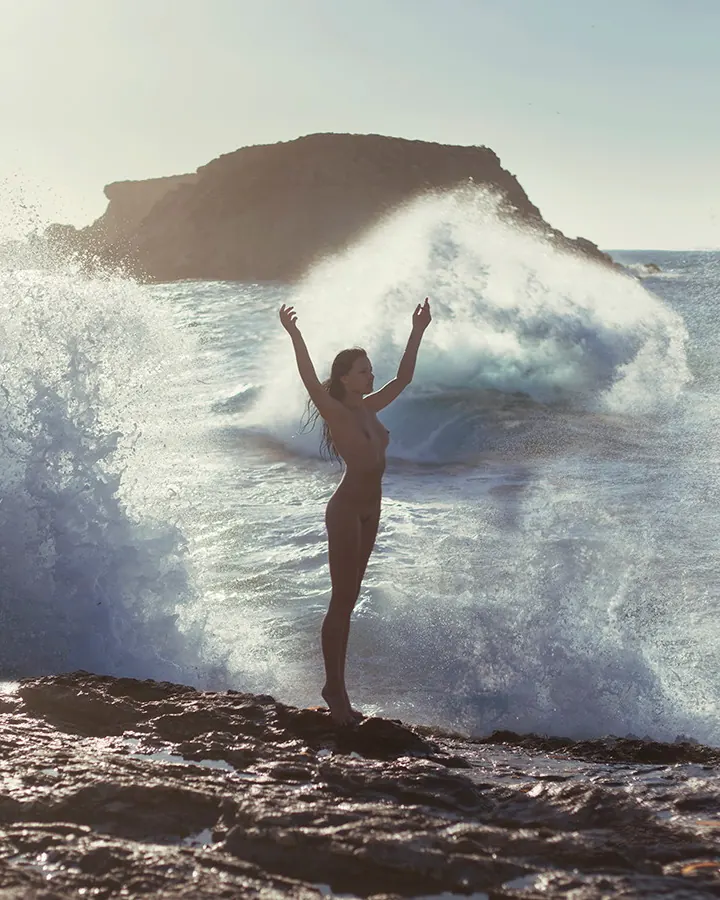 Femme nue debout sur des rochers, bras levés, devant une vague puissante.